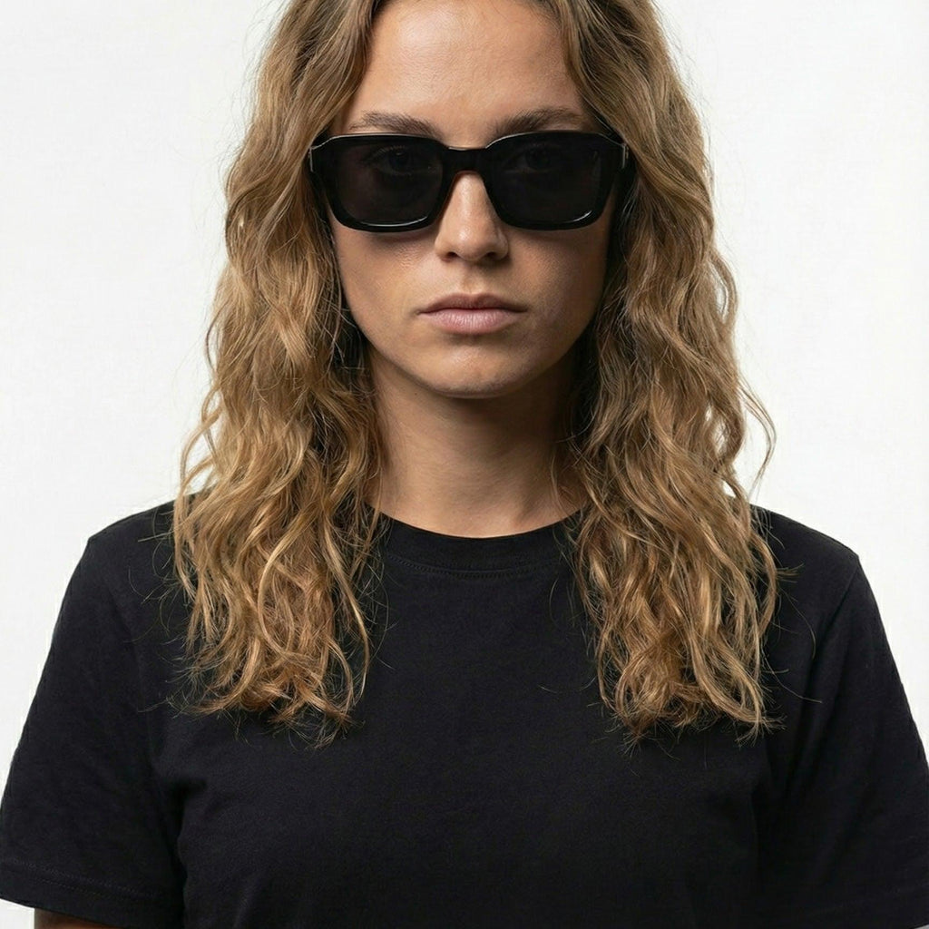 Woman with wavy hair wearing black square sunglasses with dark lenses.